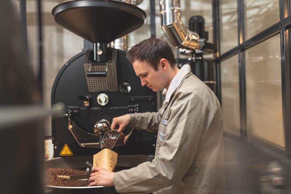 Mitarbeiter füllt Kaffeebohnen in Packung vor großem Kaffeeröster
