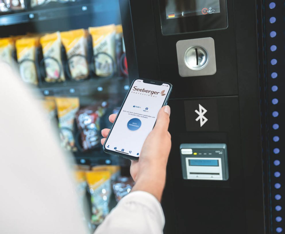 Smartphone in Hand mit geöffneter App vor Vendingautomat